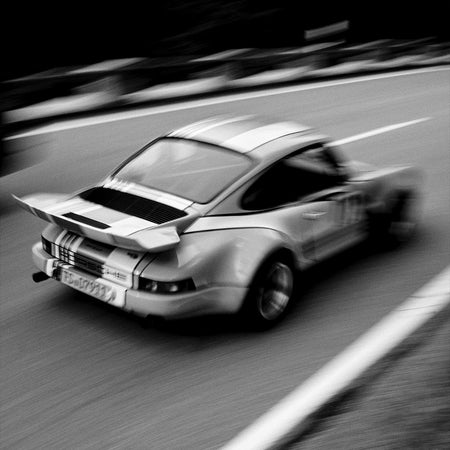 A black and white photograph of a classic sports car with wide body styling and a rear spoiler, captured in motion on a road with motion blur.