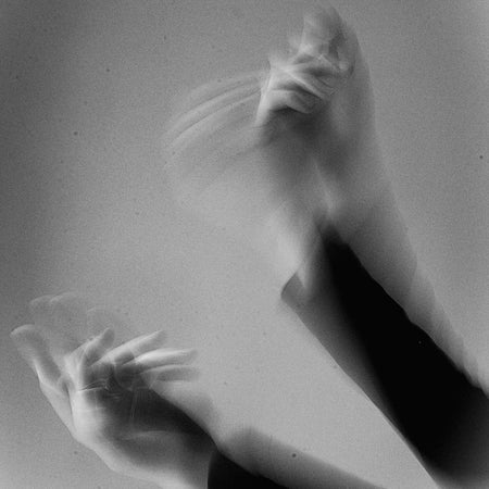 Black and white artistic photograph of two human hands and forearms in motion, creating a soft, blurred, and layered composition against a neutral gray background.