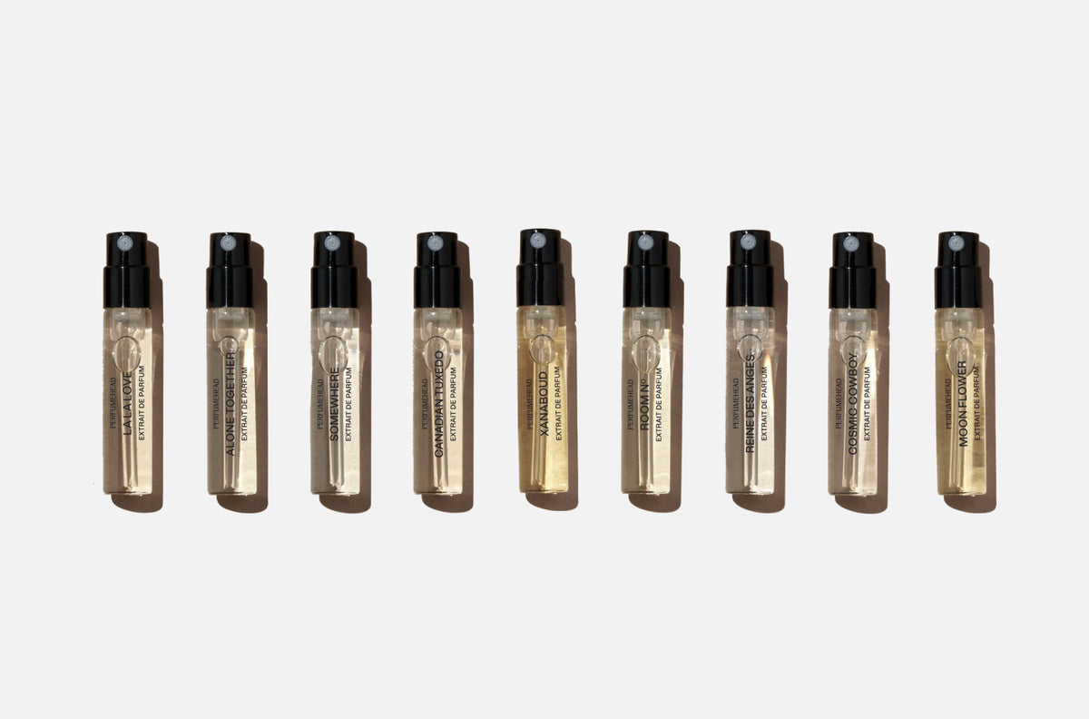 A row of nine small clear glass perfume sample vials with black spray tops, arranged in a horizontal line on a light gray background. 