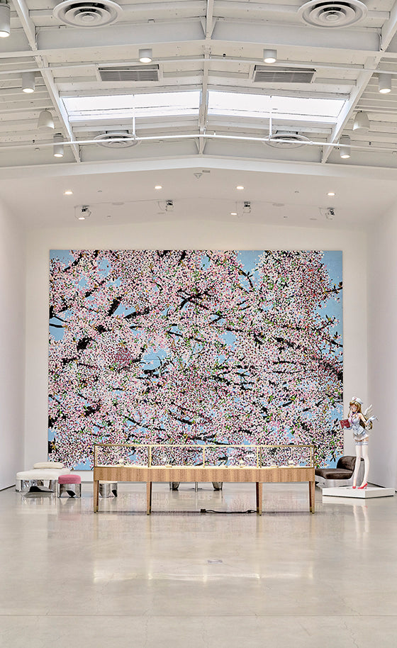 A large wall mural depicting a blooming cherry blossom tree in pink and white against a light blue sky, displayed in a modern gallery space with a wooden bench in front.