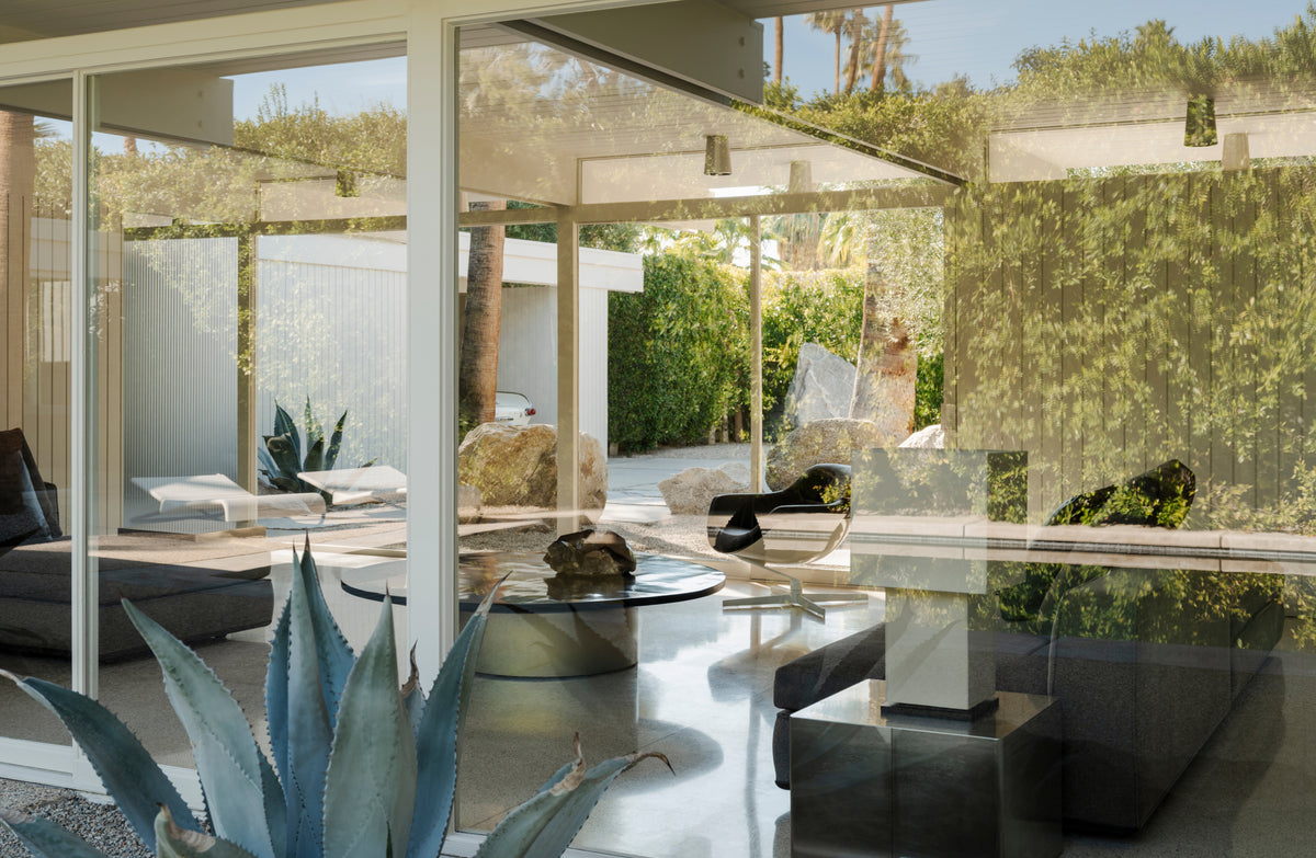 Modern glass-walled pavilion interior with outdoor seating, a round glass coffee table, and a large agave plant in the foreground, set against a lush green garden.