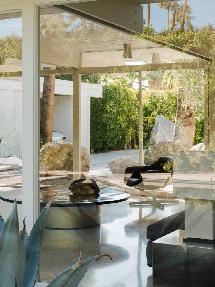 A modern interior scene with large floor-to-ceiling glass walls, a glass-top round table, a black curved lounge chair, and an outdoor patio area visible through the glass. The setting includes greenery, rocks, and palm trees, with reflections and light passing through the windows.