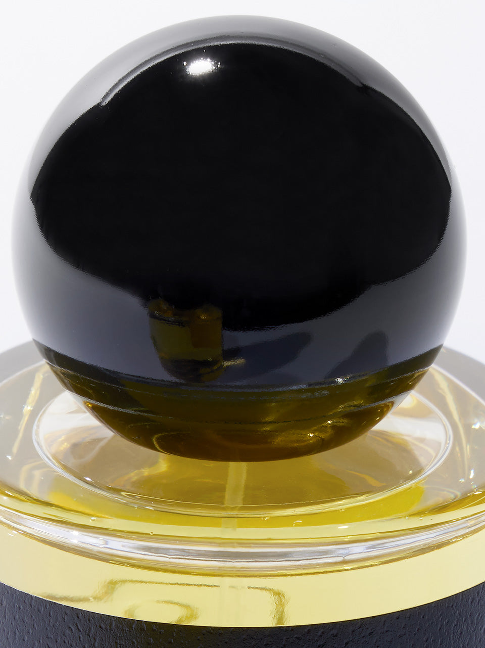 A close-up of a glossy black spherical lid resting on a glass perfume bottle with gold-colored liquid, photographed against a white background.