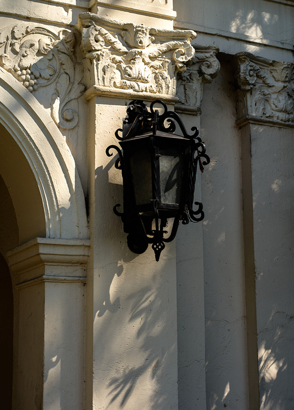 A black wrought iron wall lantern mounted on a light beige stucco building facade with ornate carved stone details and dappled shadows.