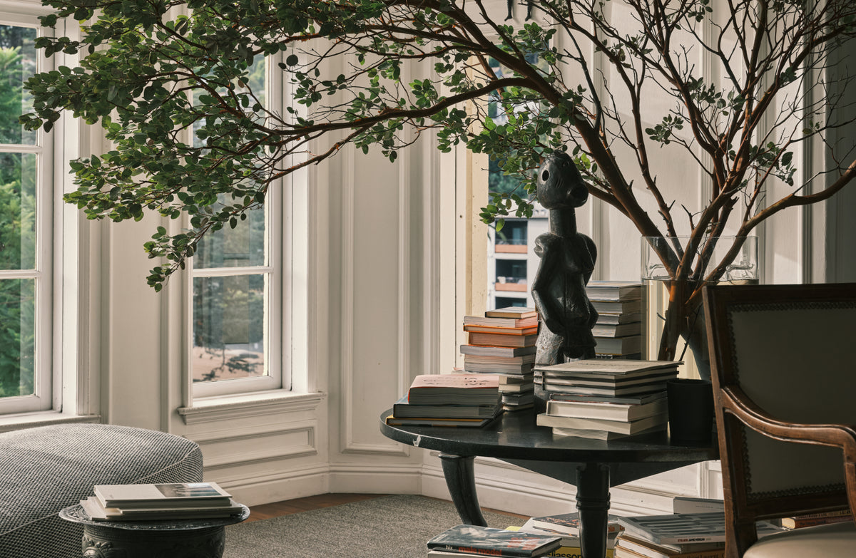 A styled interior corner with tall windows, a black round table stacked with books, a dark sculptural figurine, a leafy tree branch arrangement, and part of a gray upholstered chair.