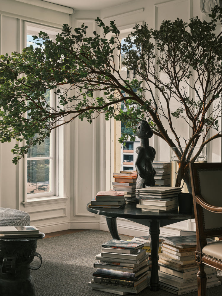 A large artificial potted tree with green foliage and brown branches displayed in a bright, classic room with white walls, windows, and a dark round table stacked with books.