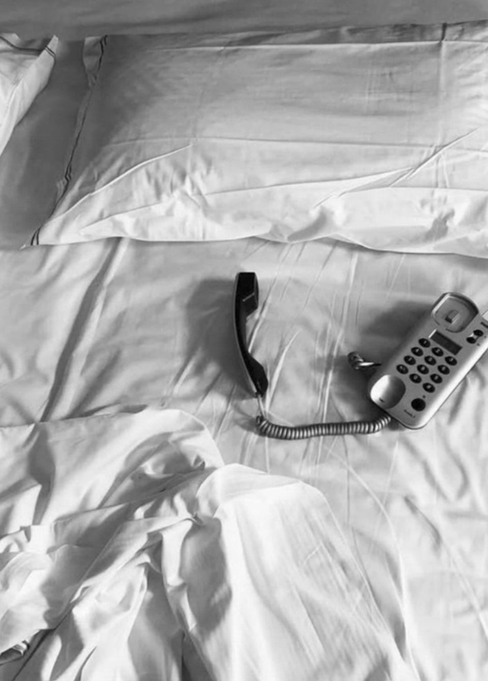 A black and white photograph of a corded landline telephone resting on rumpled white bed sheets with a pillow in the background.