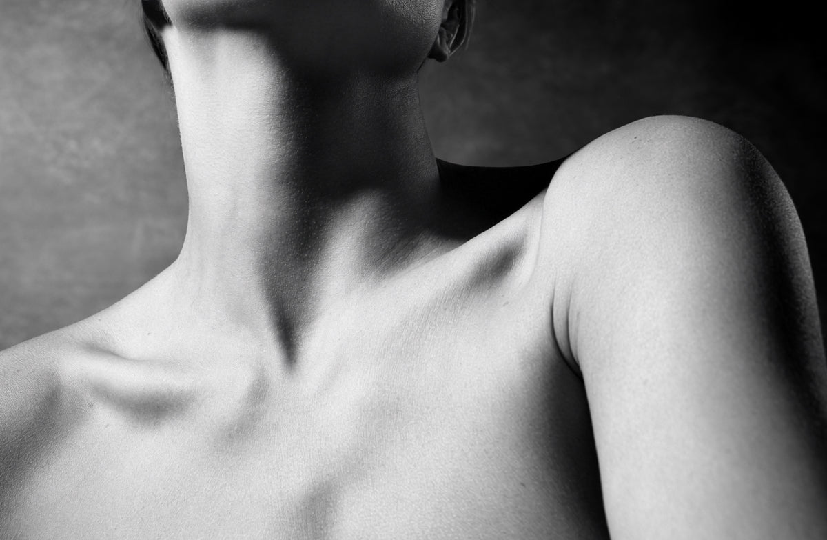 Black and white close-up photograph of a person's neck, shoulder, and upper chest against a dark studio background.