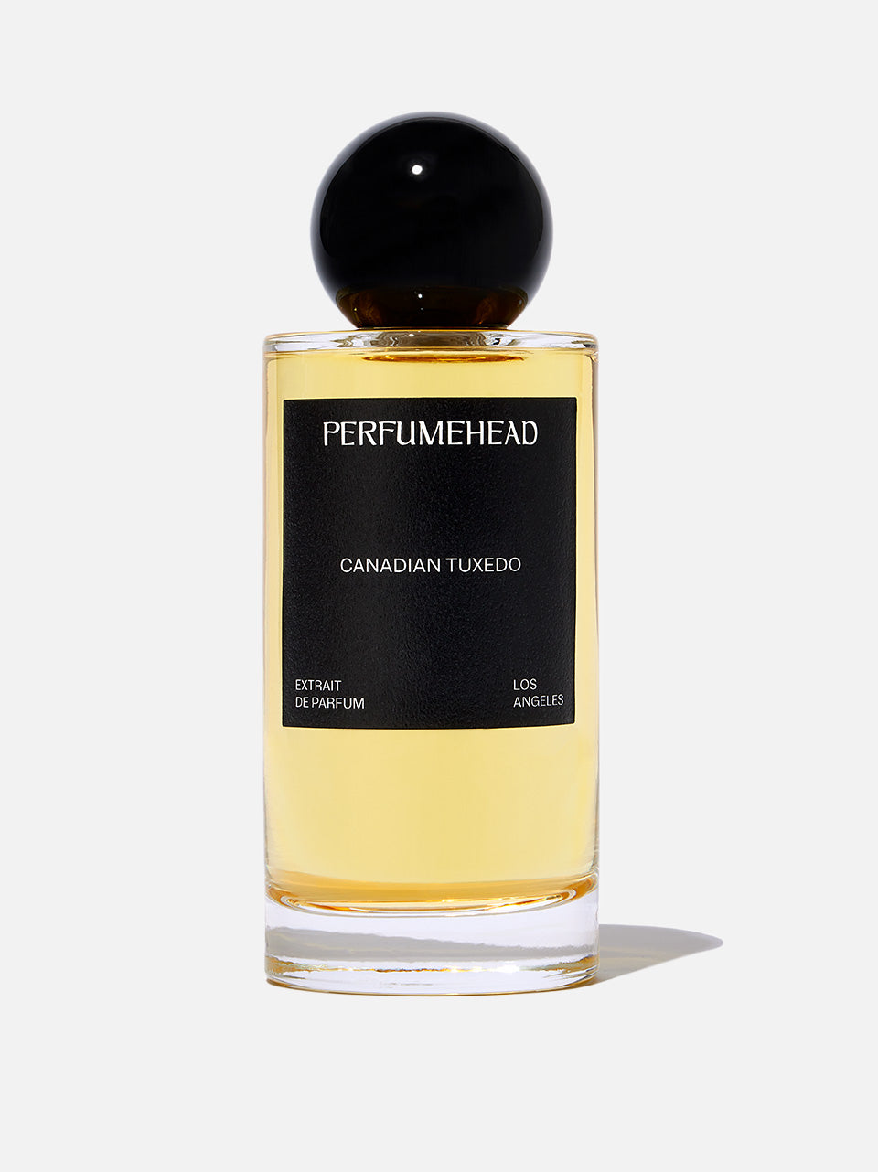 A bottle of Perfumehead 'CANADIAN TUXEDO' extrait de parfum, featuring a clear glass bottle filled with pale yellow liquid, a black label with white text, and a spherical black cap, set against a white background.