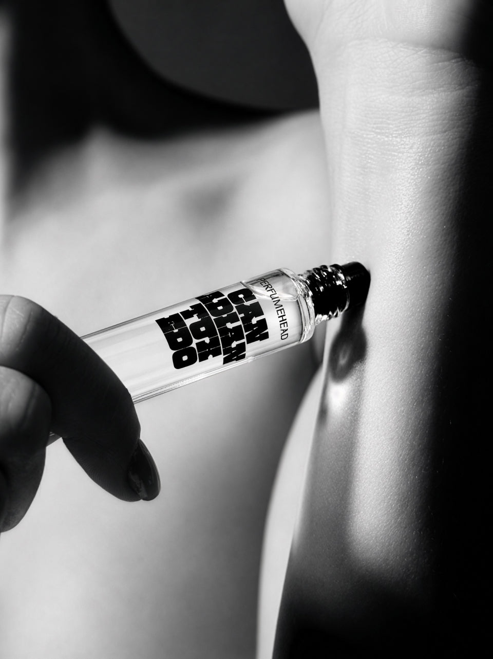 A close-up black and white photo shows a hand applying a perfume roller bottle onto a person's arm. The bottle label reads 'CANADIAN TUXEDO'.