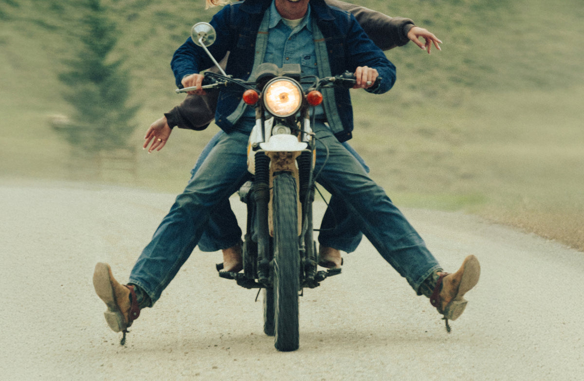 Two people riding a motorcycle on a dirt road, with the front rider wearing a blue denim jacket and jeans and the rear rider wearing a dark jacket and jeans. The motorcycle has a round headlight and a vintage aesthetic.