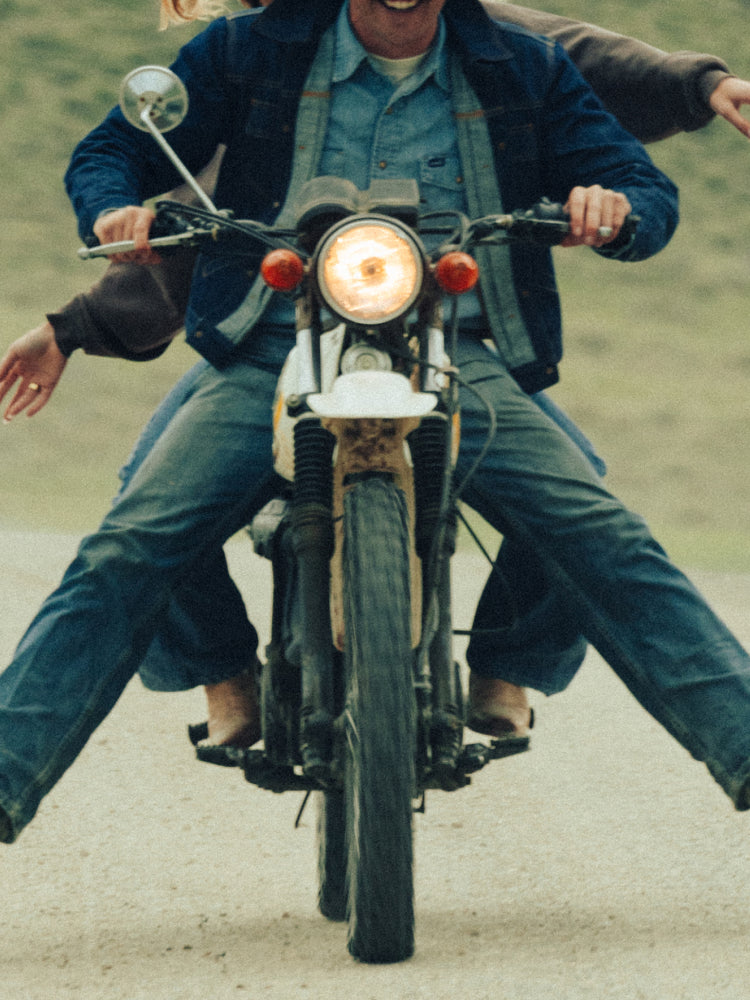 A person riding a vintage-style motorcycle with another person seated behind them, photographed outdoors on a dirt road. The riders are wearing blue denim clothing, and the motorcycle has a round headlight, red turn signals, and a cream-colored front fender.