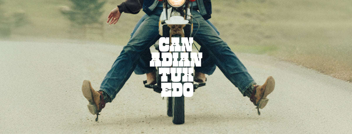 A person riding a motorcycle on an open road with legs extended outward in a wheelie pose. The rider is wearing blue jeans and brown boots, and the motorcycle has a round headlight and a visible front wheel. White stylized text in the centre saying 'CANADIAN TUXEDO'. 