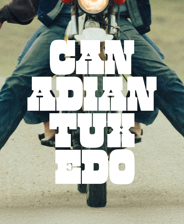 A stylized graphic with large white block text reading 'CANADIAN TUXEDO' overlaid on a photo of a person riding a motorcycle, cropped from the back, with blue jeans and a dark jacket visible against a muted beige-gray background.