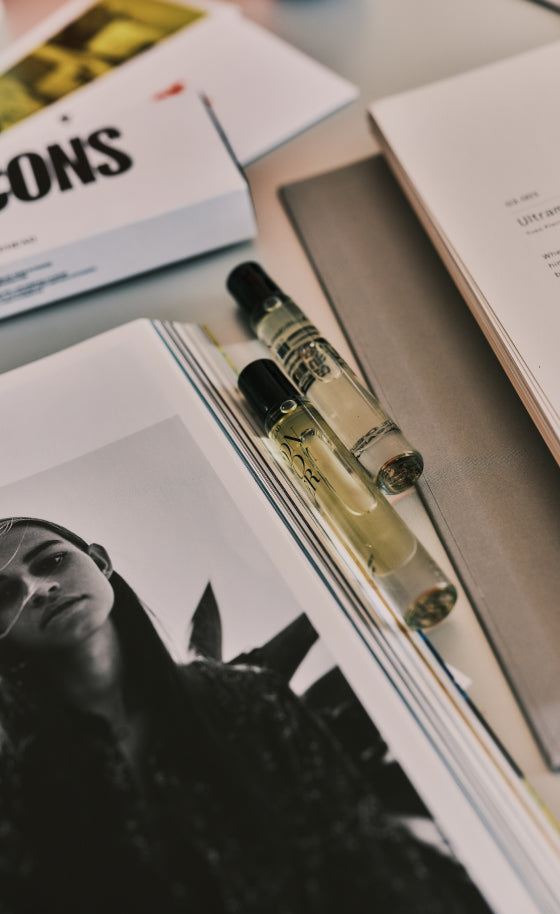 Two clear glass perfume sample vials with black caps resting on an open book, surrounded by other books on a tabletop.
