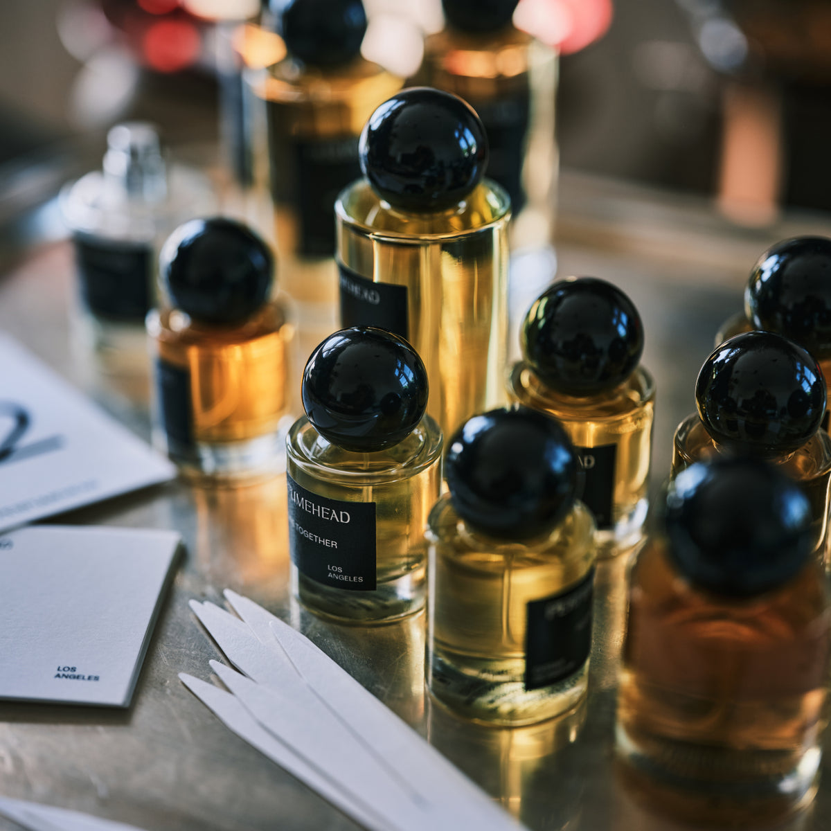 A group of amber glass perfume bottles with spherical black caps arranged on a reflective metallic surface, with a few white cards in the foreground.