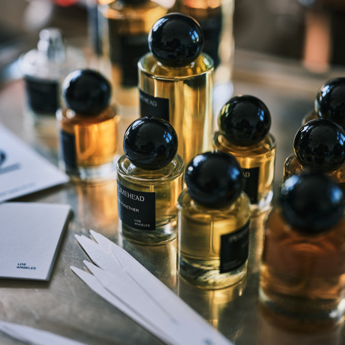 A close-up, shallow depth-of-field shot of several amber glass perfume bottles with black spherical caps and black labels, arranged on a reflective metallic surface with white paper cards nearby.