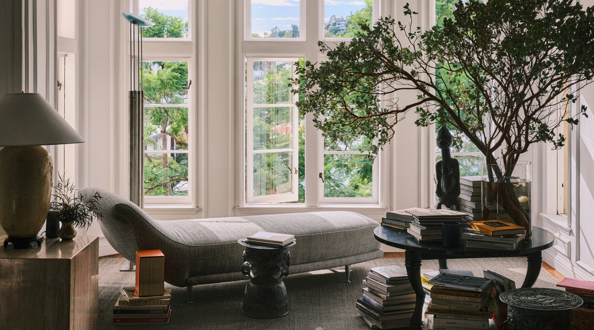 A styled interior corner with tall white-framed windows, a gray upholstered chaise lounge, a black side table stacked with books, a floor lamp, a large potted tree, and outdoor greenery visible in the background.