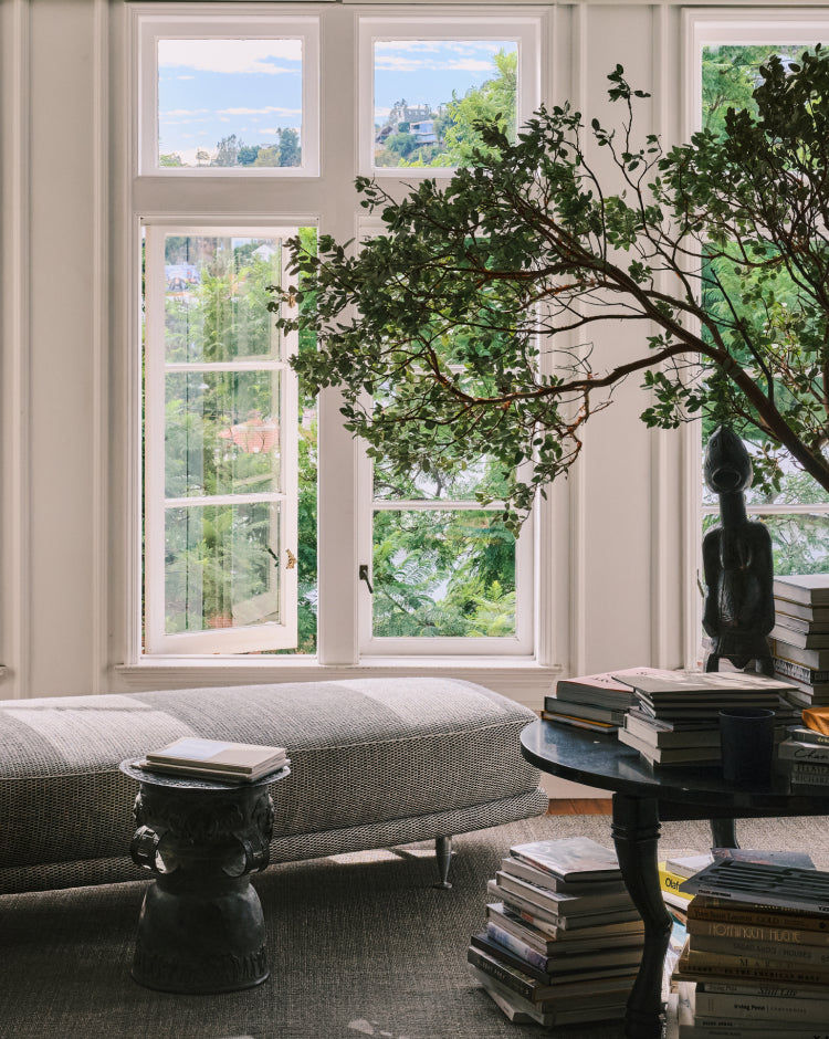 A styled interior scene with a large gray upholstered bench or chaise against tall white-framed windows, a dark round table stacked with books, a sculptural statue, and a leafy tree branch in the foreground. 
