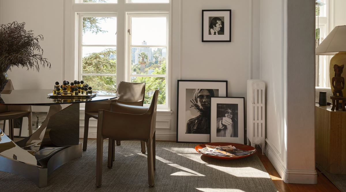 A styled interior room with a glass-top dining table, brown upholstered chairs, framed black-and-white wall art, and three larger framed prints leaning against a white wall near a window.