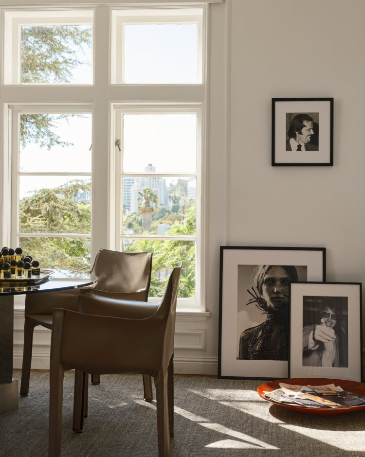 A styled interior scene with a round dining table, two brown leather chairs, framed black-and-white portraits on a white wall, and a tray of magazines on a carpeted floor in a sunlit room.