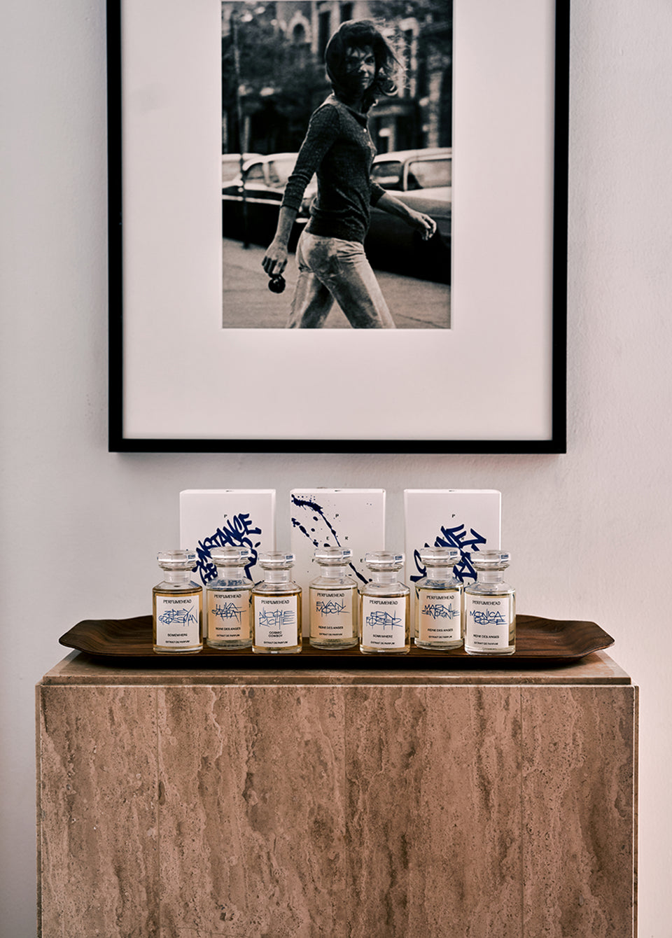 A styled interior scene showing a framed black-and-white street photograph hanging on a light wall above a stone pedestal. On the pedestal sits a dark wooden tray holding several small glass jars with labels and white boxes featuring blue abstract designs.