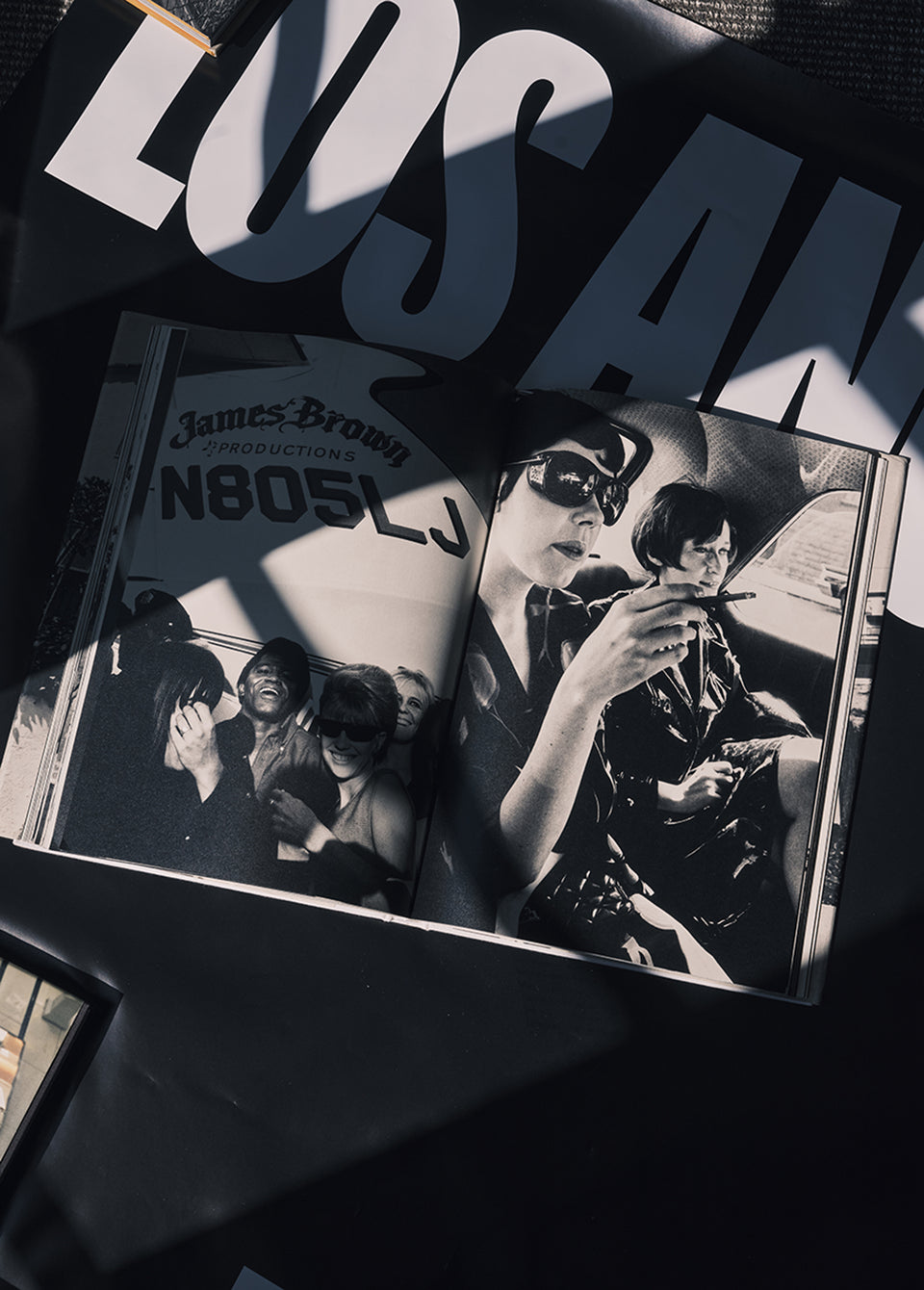 An open black-and-white photo book lies on a dark surface under dramatic diagonal shadows. The pages show black-and-white portraits and text, including 'James Brown Productions N805LJ' and large partial letters 'LOS AN.