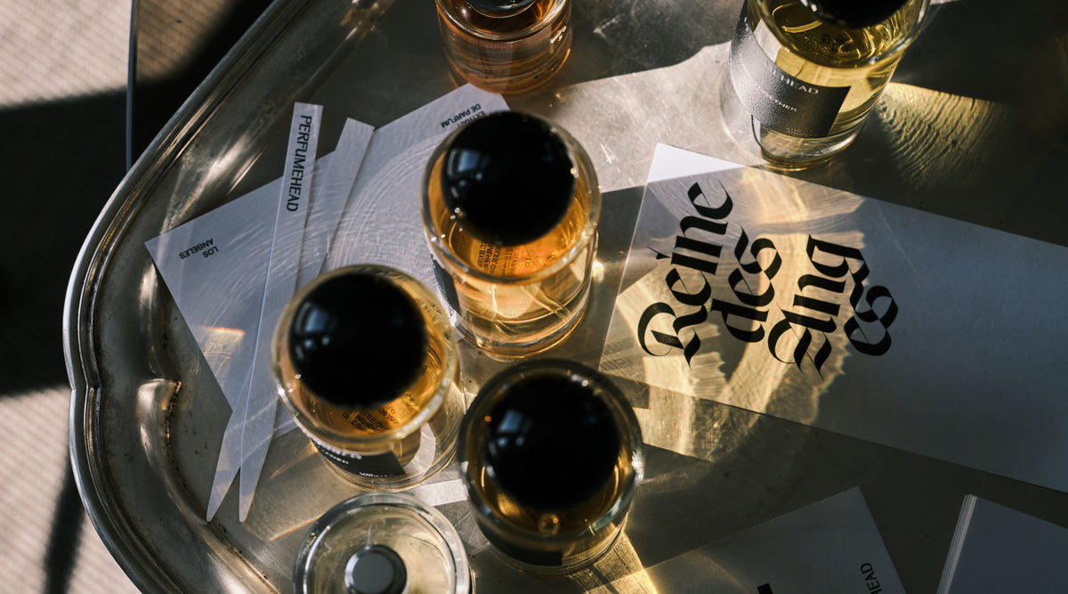 Several clear glass perfume bottles with black caps containing amber liquid, arranged on a reflective silver tray with printed cards under warm sunlight.