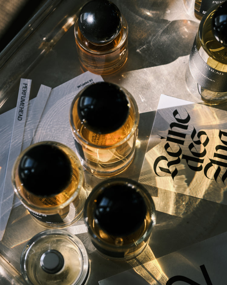 Several perfume bottles with black spherical caps and amber liquid are arranged on reflective metallic paper and white cards. One card shows the text 'PERFUMEHEAD' and another has large gothic lettering.