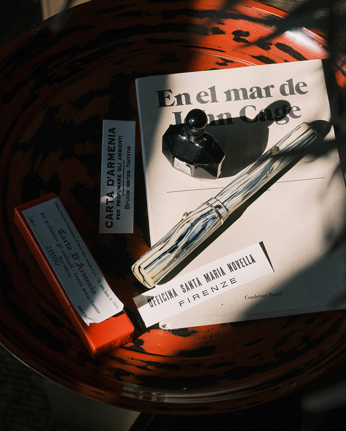A styled flat-lay featuring a vintage striped fountain pen resting on a book titled 'En el mar de John Cage', alongside a small orange box, a white card labeled 'Carta d'Armienia', and a black glass object inside a dark orange marbled bowl.