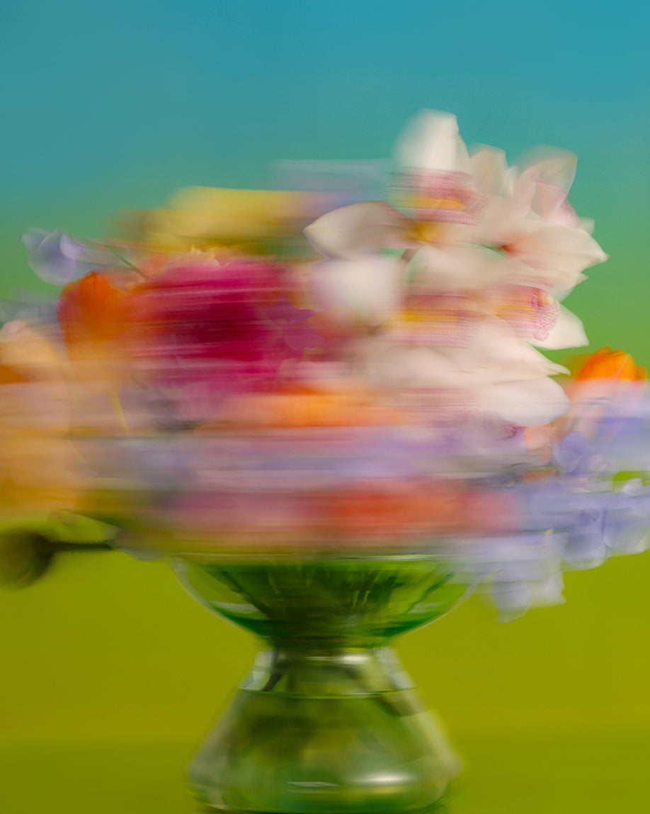 A blurred bouquet of multicolored flowers in a clear glass pedestal bowl, photographed against a bright green and blue gradient background.