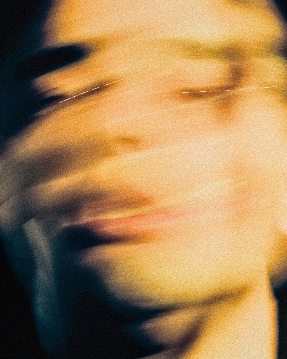 An abstract, motion-blurred portrait of a person's face with soft warm yellow and orange tones against a dark background.