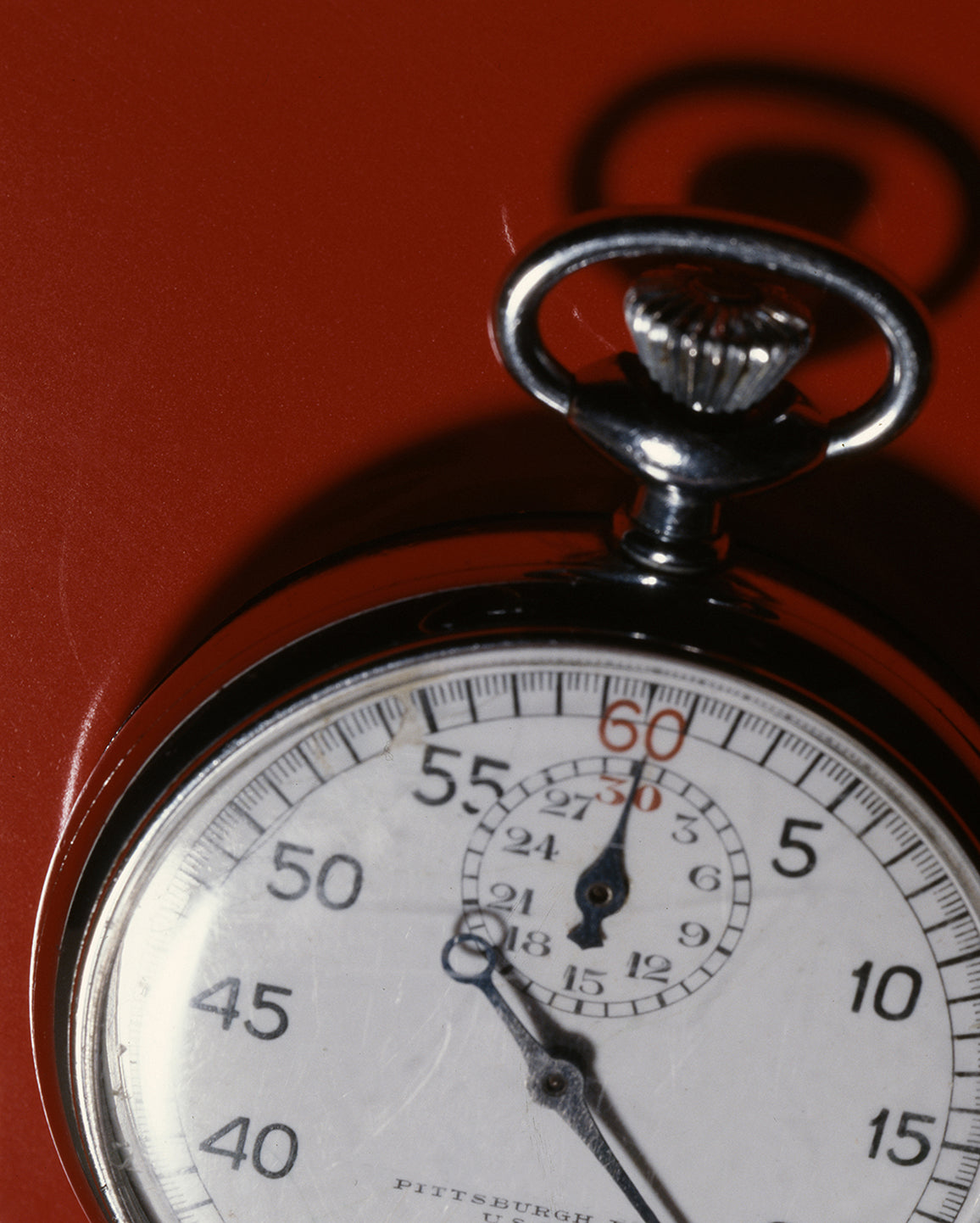 A close-up shot of a vintage pocket watch with a silver-toned metal case and white dial, featuring black numerals and red second hand markings, set against a solid red background.
