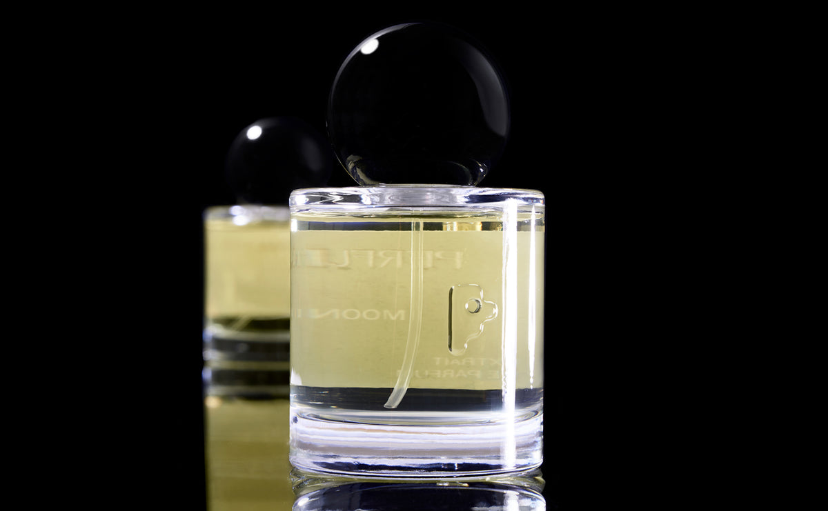 A clear glass perfume bottle with pale yellow liquid and a glossy black spherical cap, shown in front of a matching bottle on a black reflective background.