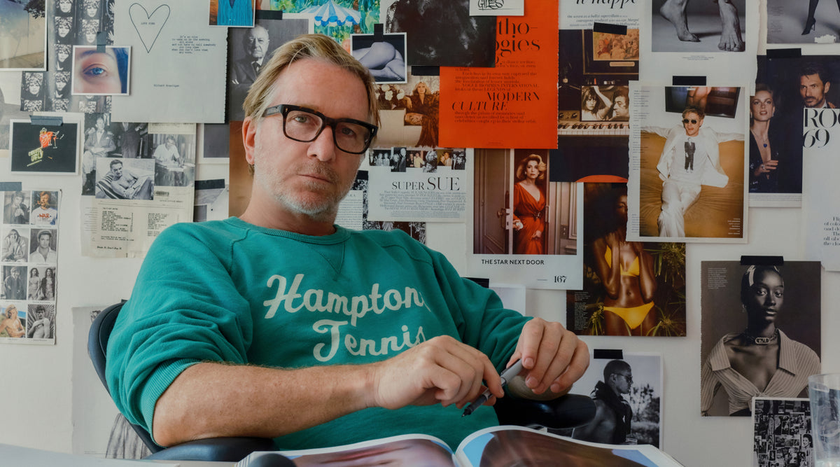 A man with glasses and a beard wearing a green long-sleeve shirt with 'Hampton Tennis' printed on the front, sitting in front of a wall covered in posters and art prints.