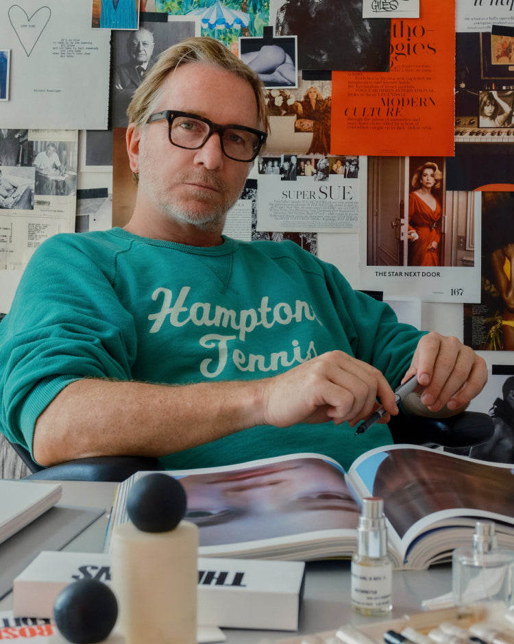 A man wearing black-rimmed glasses and a green long-sleeve sweatshirt with 'Hampton Tennis' printed in white across the chest, seated at a desk with books, perfume bottles, and a magazine in a creative office environment.