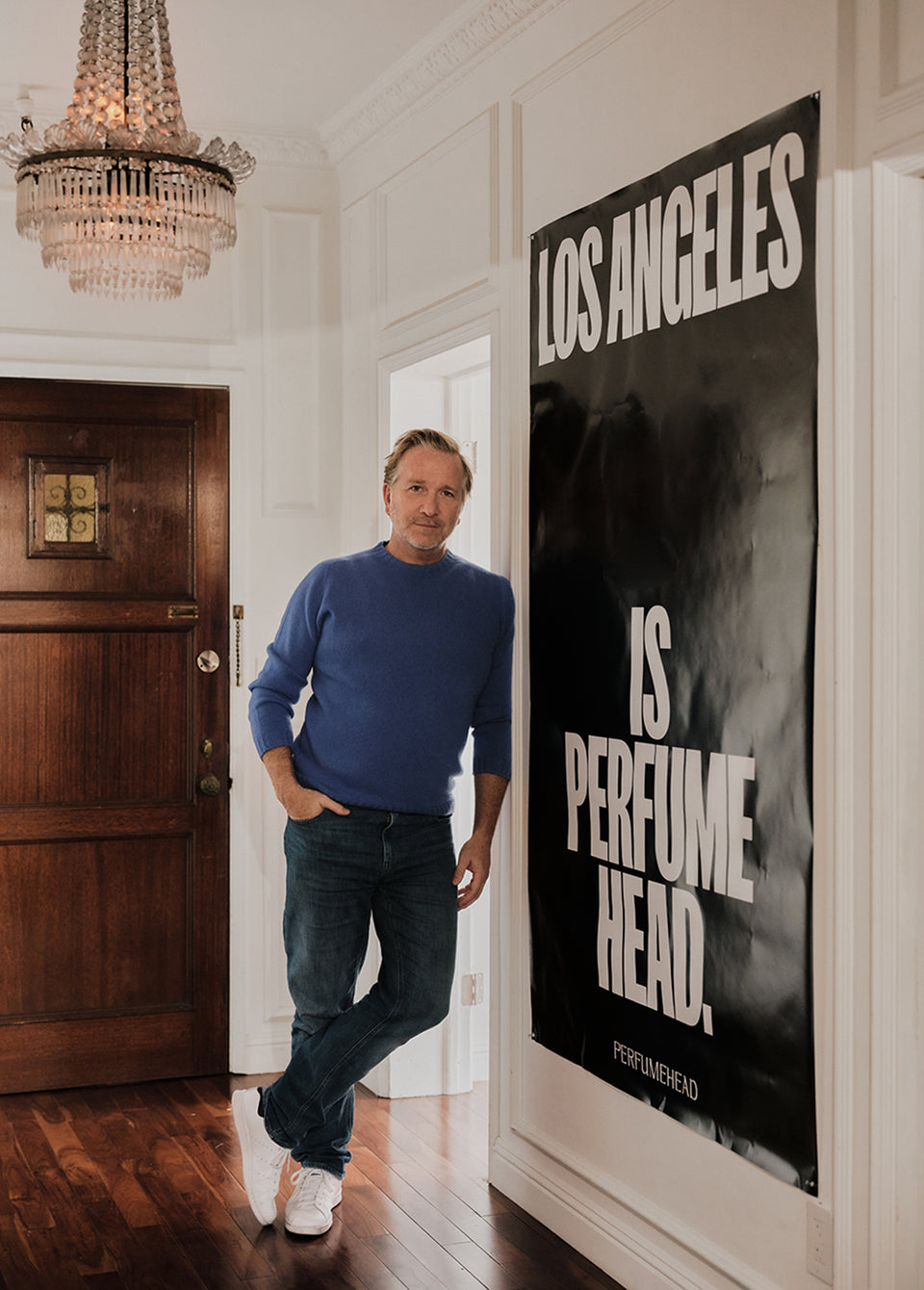A man in a blue sweater and jeans stands in a hallway next to a large black poster with white text that reads 'LOS ANGELES IS PERFUME HEAD.' A crystal chandelier hangs from the ceiling, and a dark wood door is on the left.