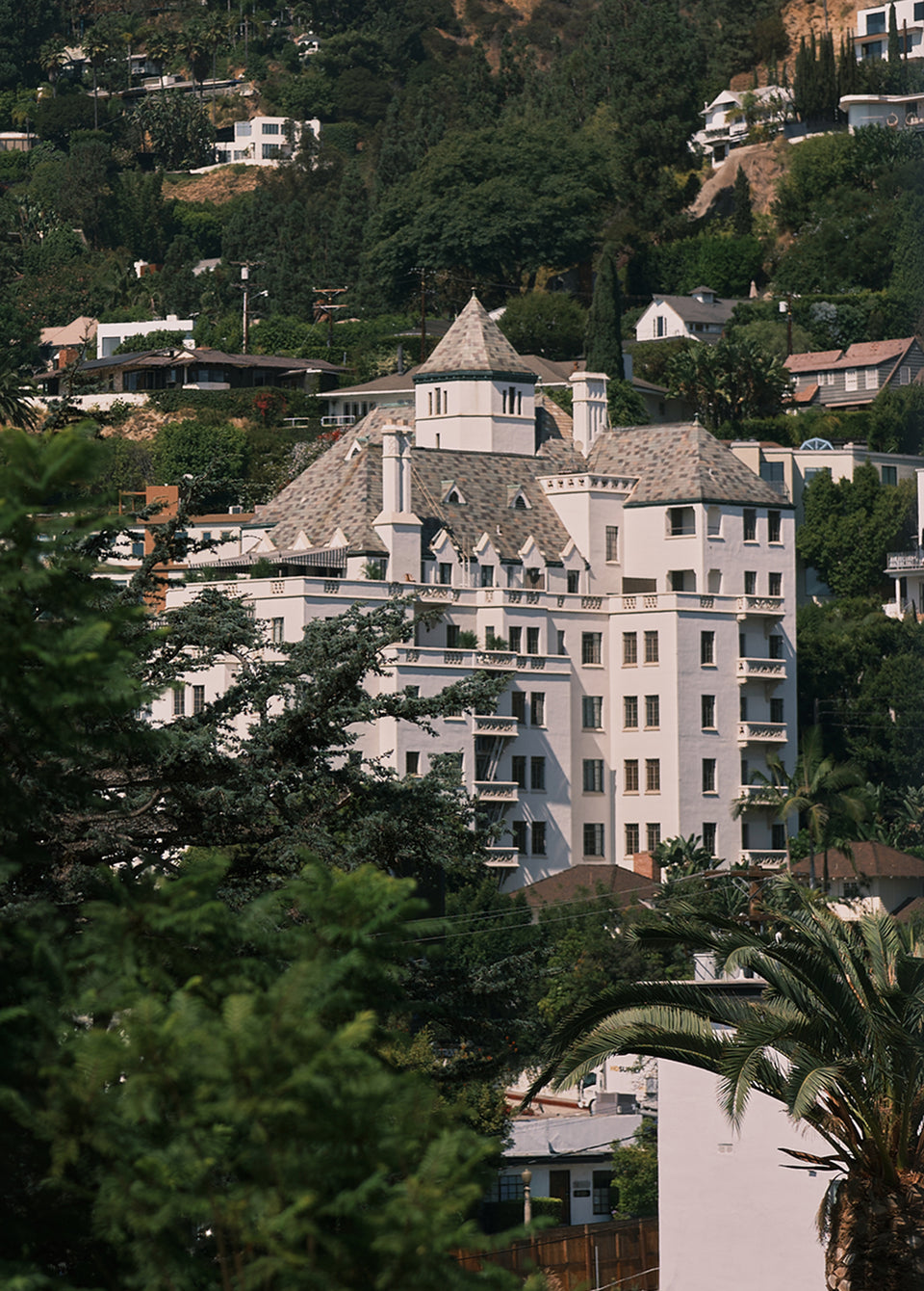 A large white historic-style mansion with gray shingled roofs, surrounded by dense green trees and hills with other houses in the background.