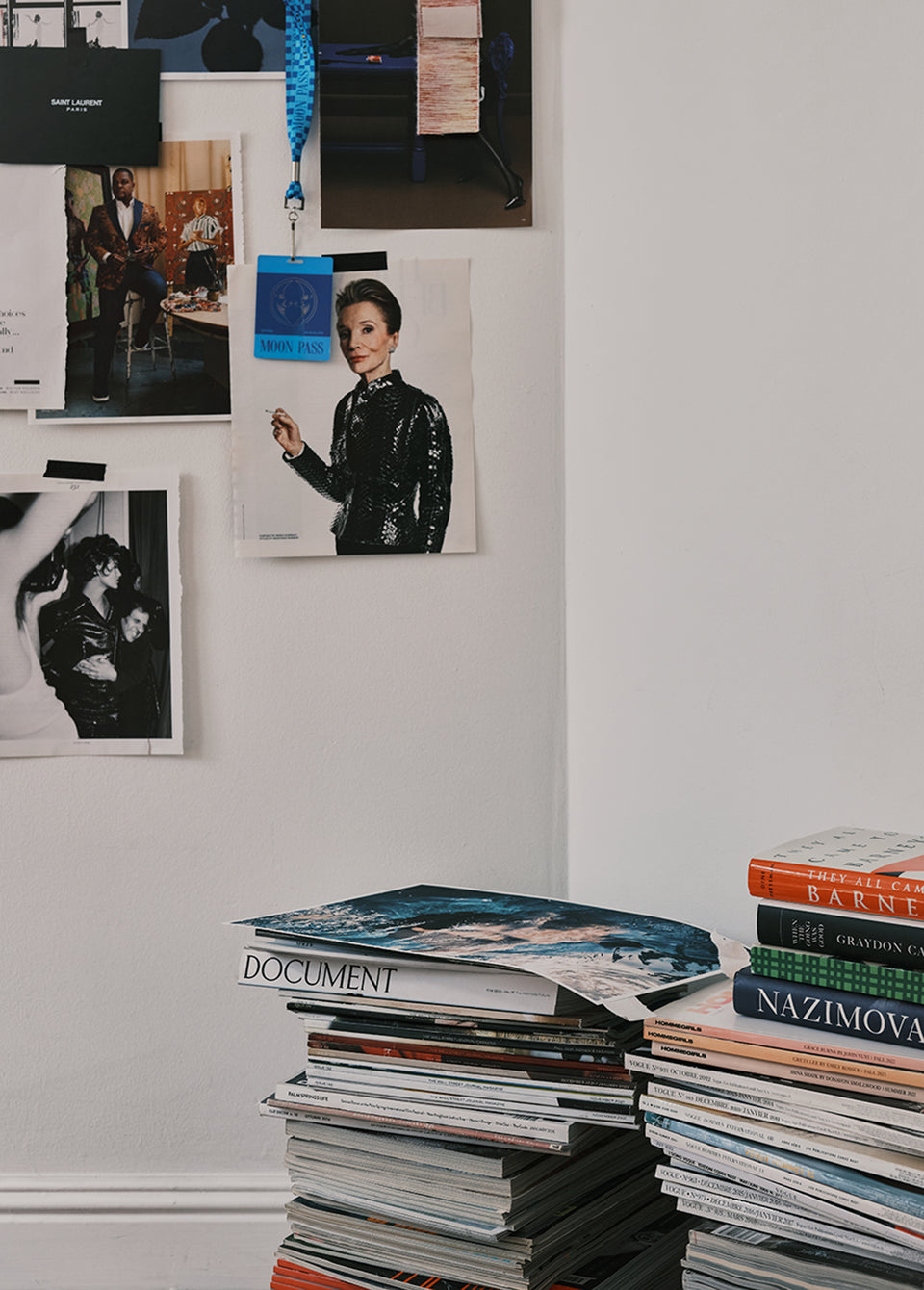 A styled interior corner with a white wall decorated with framed and pinned fashion and art prints, alongside tall stacks of magazines and books in the foreground.