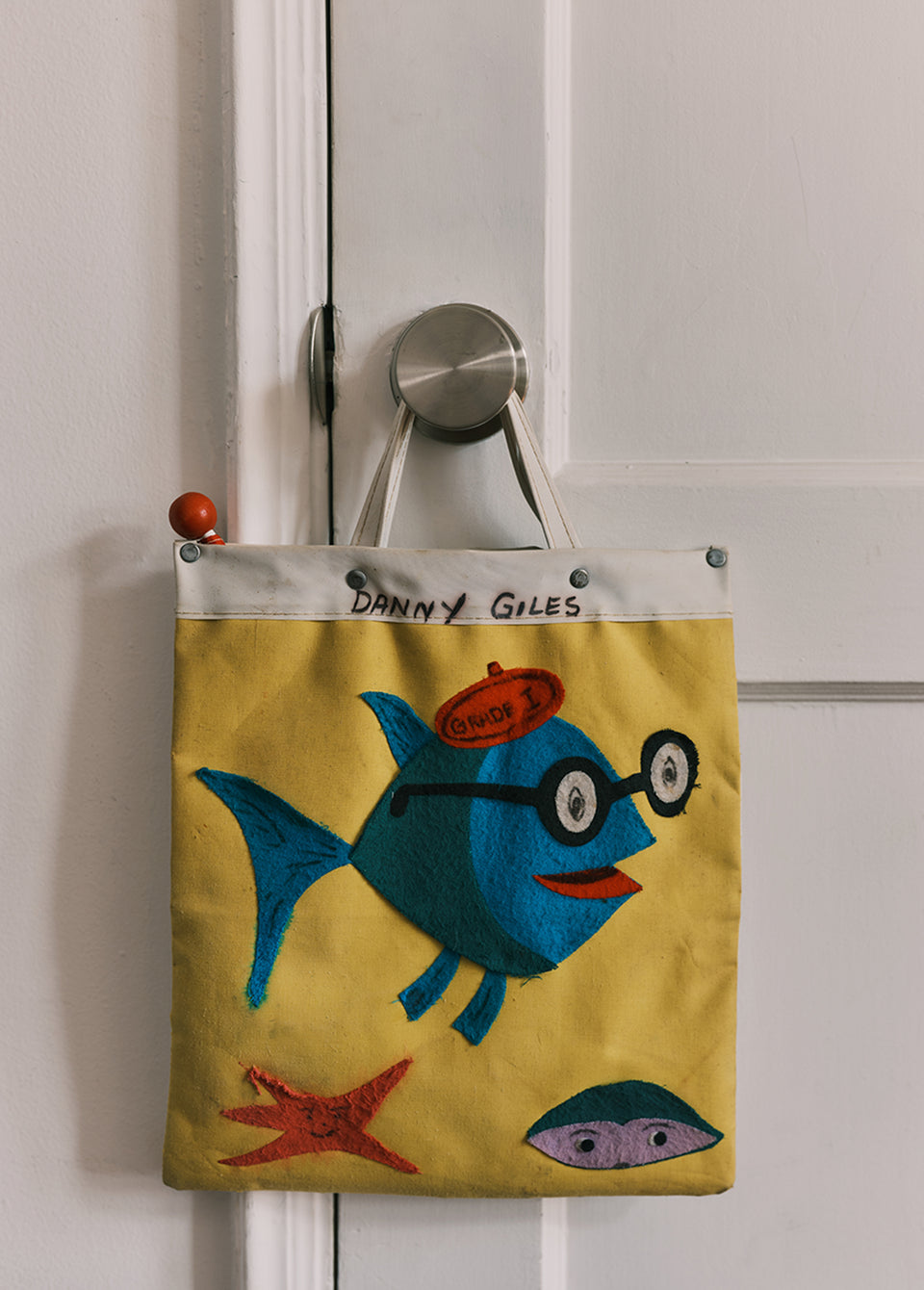 A yellow fabric tote bag with a whimsical embroidered blue fish, red starfish, and clam design, hanging from a silver door hook on a white door. The top of the bag has a white band with the text 'DANNY GILES'.