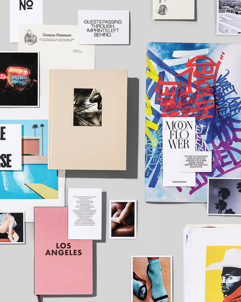 A styled flat-lay collage of various printed items, including a pink notebook with 'LOS ANGELES' in black, a cream-colored book with a black-and-white photo, a colorful magazine or poster with 'MOON FLOWER' and graffiti-style lettering, and several small cards and photos arranged on a light gray surface.