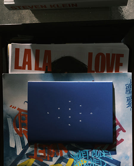 A dark blue rectangular box with small white geometric symbols on top, placed over a stack of printed materials including a white sheet with red 'LA LA LOVE' text and a blue illustrated card. A partial book or box at the top shows 'STEVEN KLEIN'.