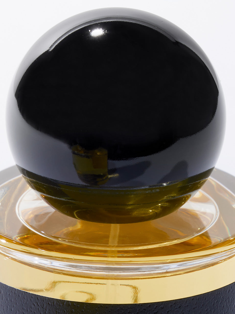 A close-up of a spherical black bottle cap resting on a clear glass base filled with amber-yellow liquid, set against a white background.