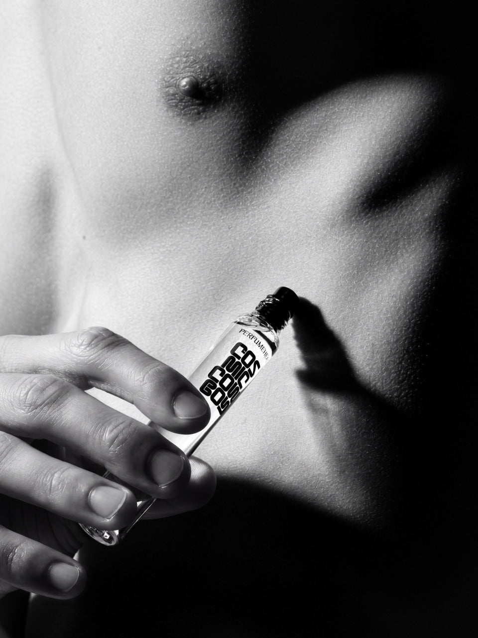 A close-up black and white photograph of a person's bare chest and hand holding a small cylindrical perfume roller near the skin.