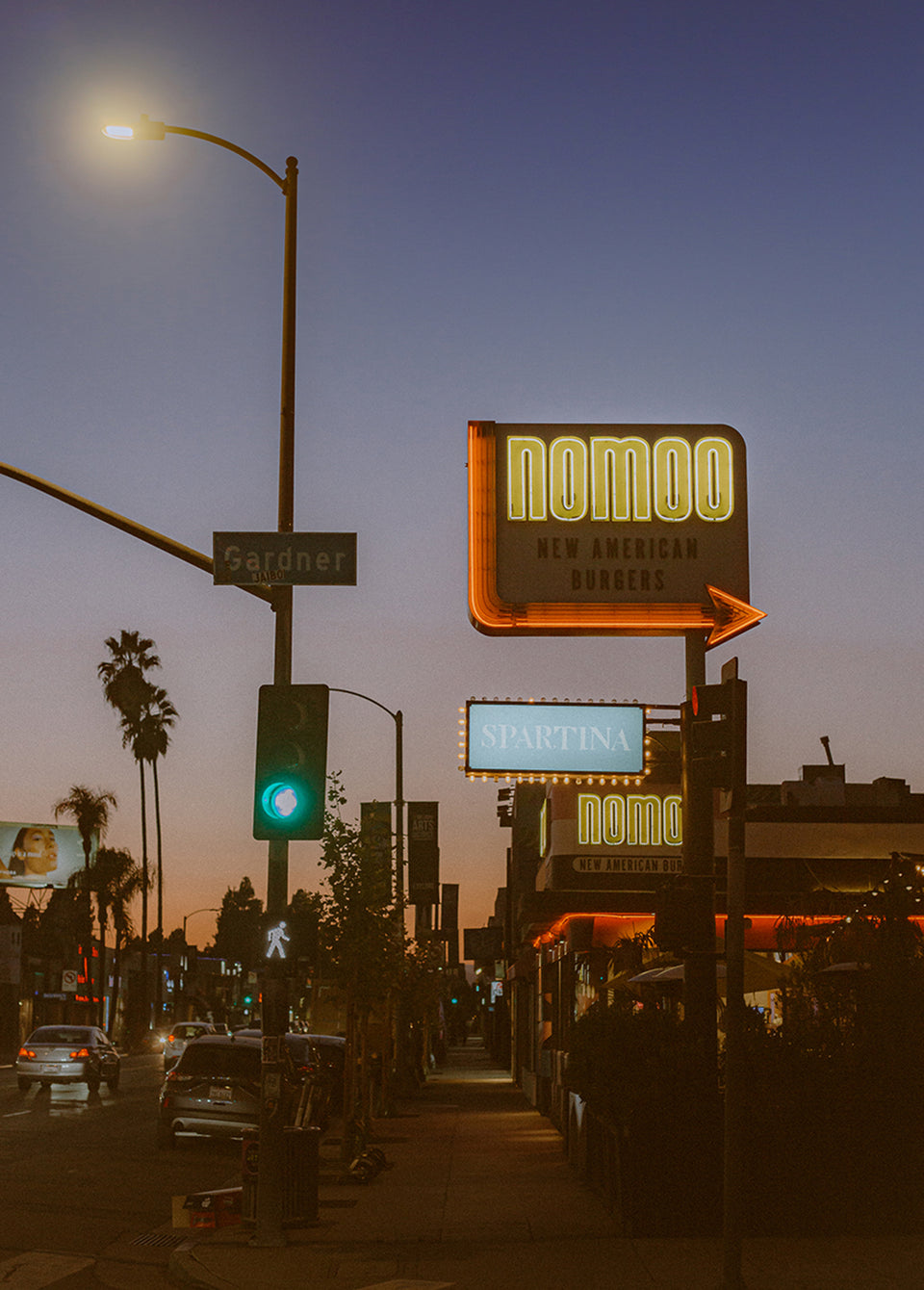 A street scene at dusk with illuminated restaurant signs, including a prominent 'nomoo' sign, palm trees, cars, and a street lamp against a blue twilight sky.