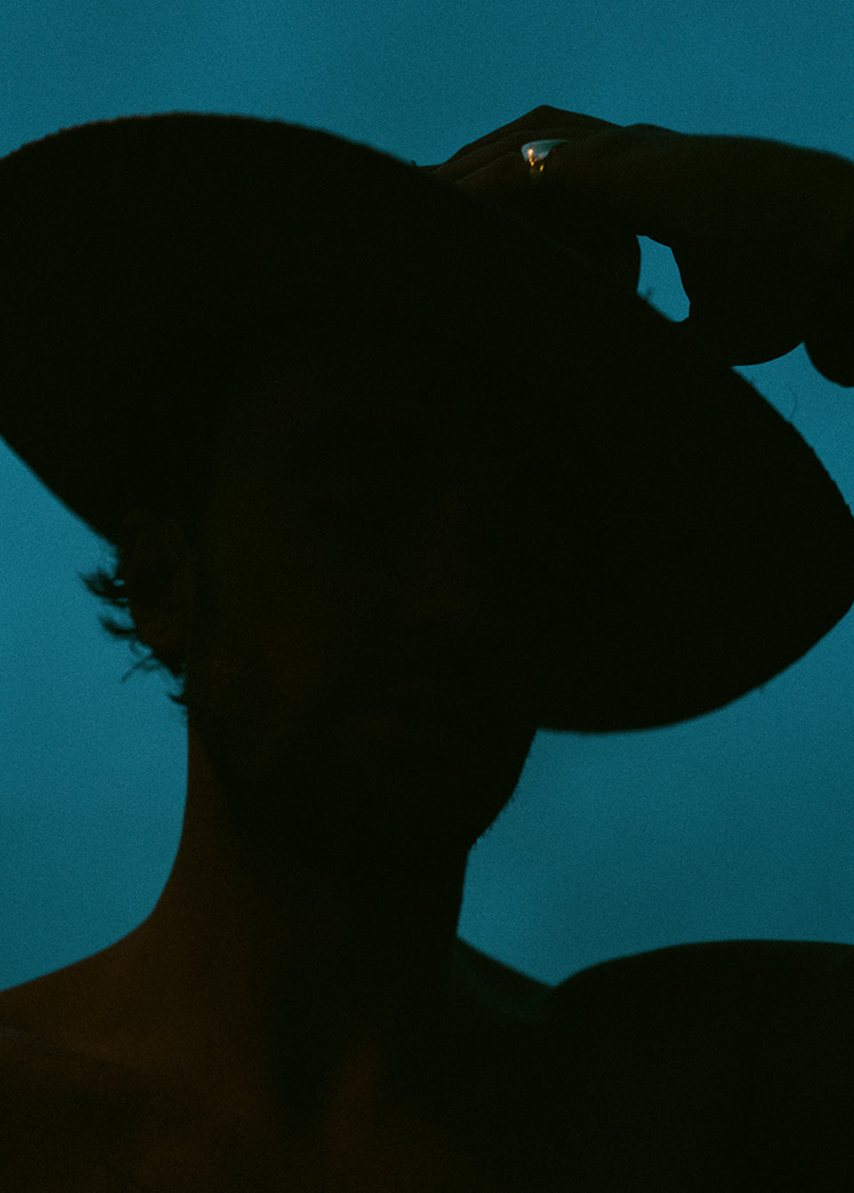A dramatic silhouette portrait of a person wearing a wide-brimmed hat against a deep teal-blue background.