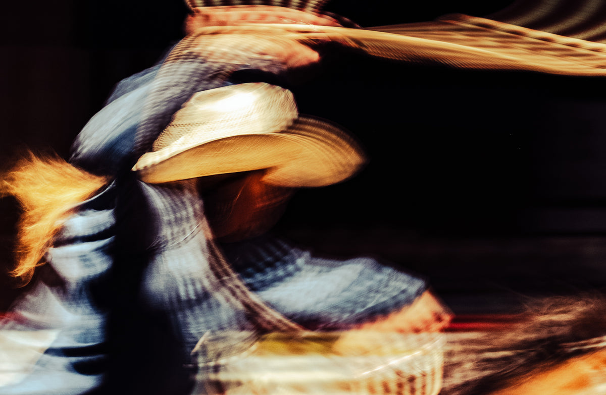 A motion-blurred close-up of a person wearing a light-colored cowboy hat, with warm orange highlights against a dark background.