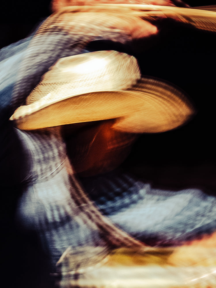 A close-up, motion-blurred photograph of a person wearing a light beige cowboy hat and a blue plaid shirt against a dark background.