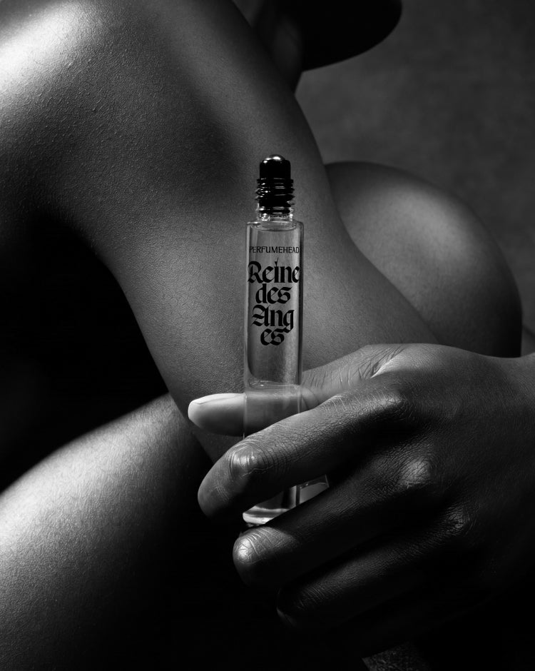 A black and white close-up photo of a person holding a clear glass perfume rollerball bottle labeled 'Reine des Anges' against their skin.