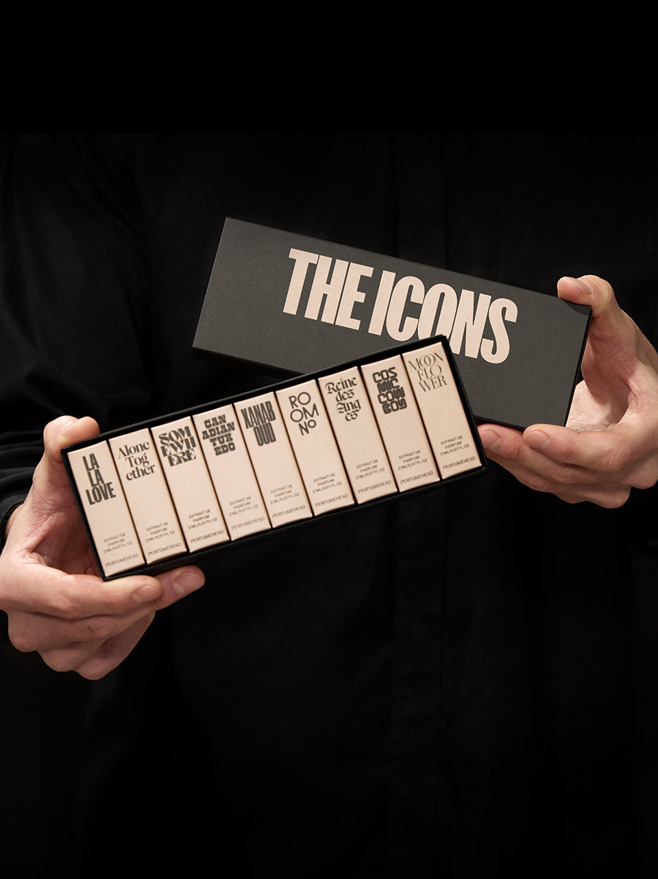 A person in a black shirt holds a set of nine rectangular perfume boxes in a larger box labeled 'THE ICONS'. The boxes are cream with black text, and the person is shown against a black background.